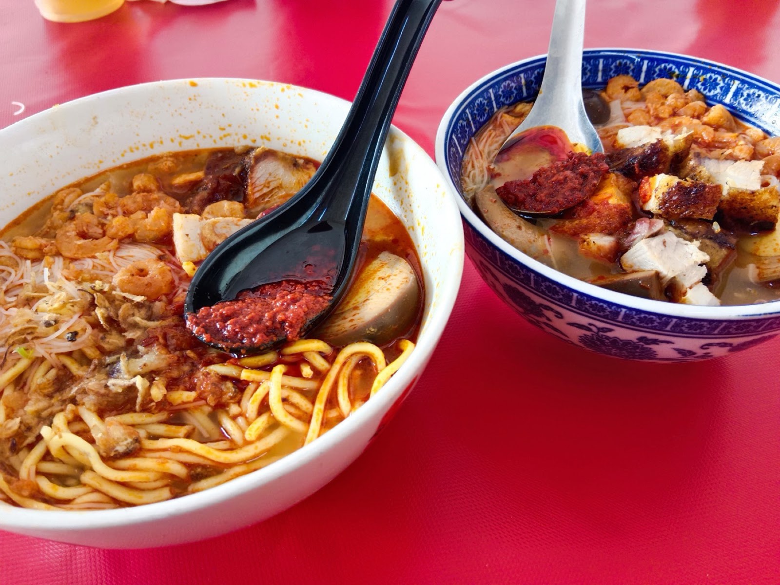 Two bowls of Hokkien prawn mee in a red prawn-and-pork broth with sambal, prawns, pork slices and yellow noodles on a red table