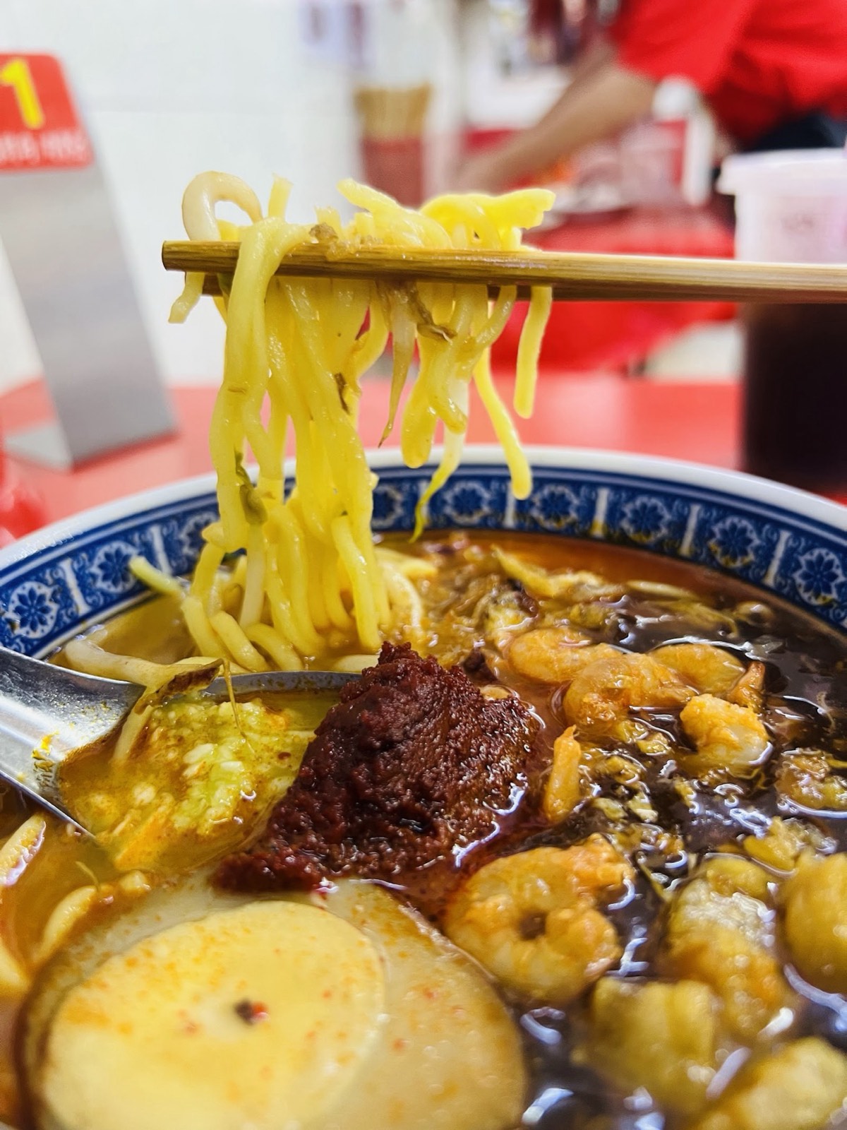 Chopsticks lifting springy yellow Hokkien noodles from a bowl of dark prawn broth, a blob of red chili sambal beside fresh prawns