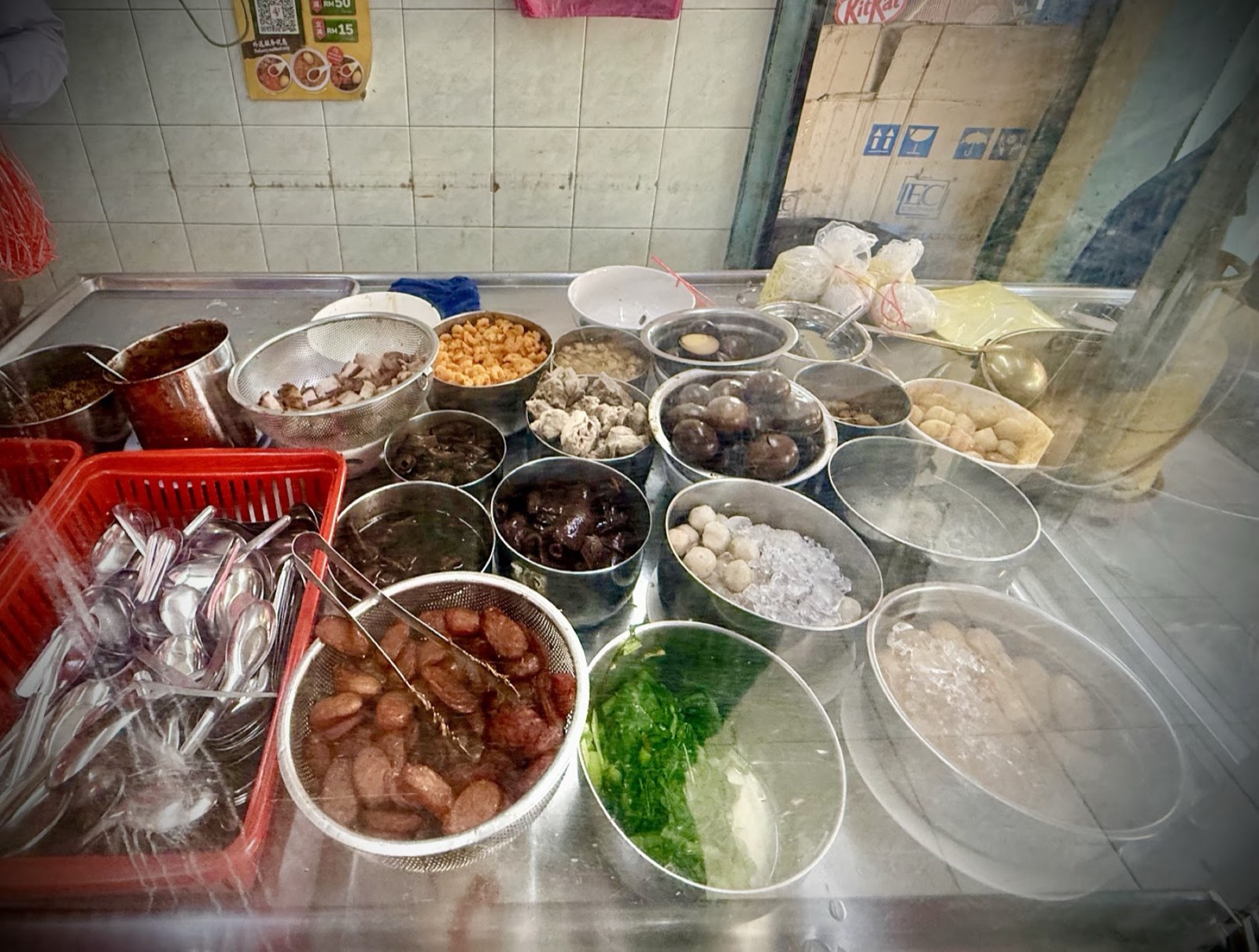 The stainless-steel toppings counter with bowls of pork, mushrooms, fishballs, braised egg and greens behind glass